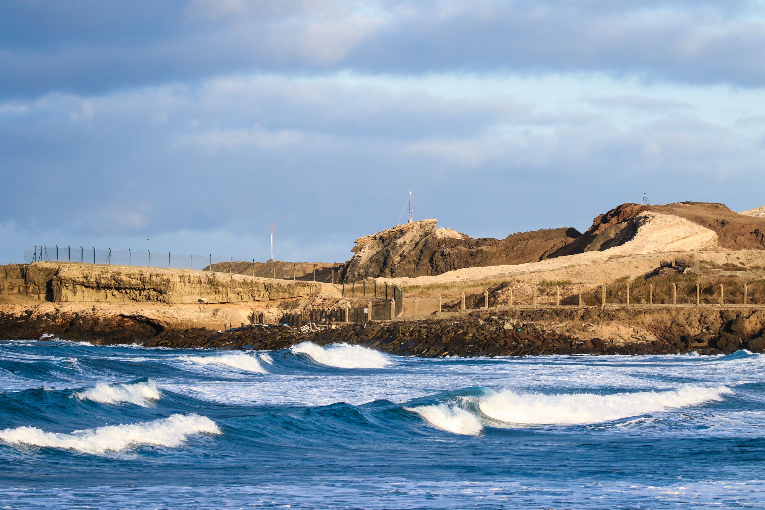 A scenic view of waves crashing on a rocky Spanish coastline under a blue sky.